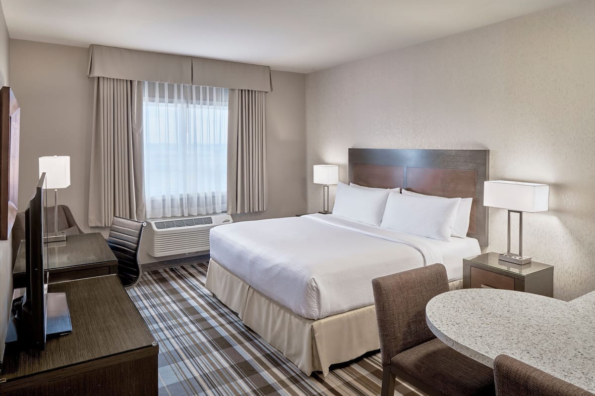 A hotel room with a bed, two bedside tables with lamps, a desk with a chair, a small round table, and a window with curtains.