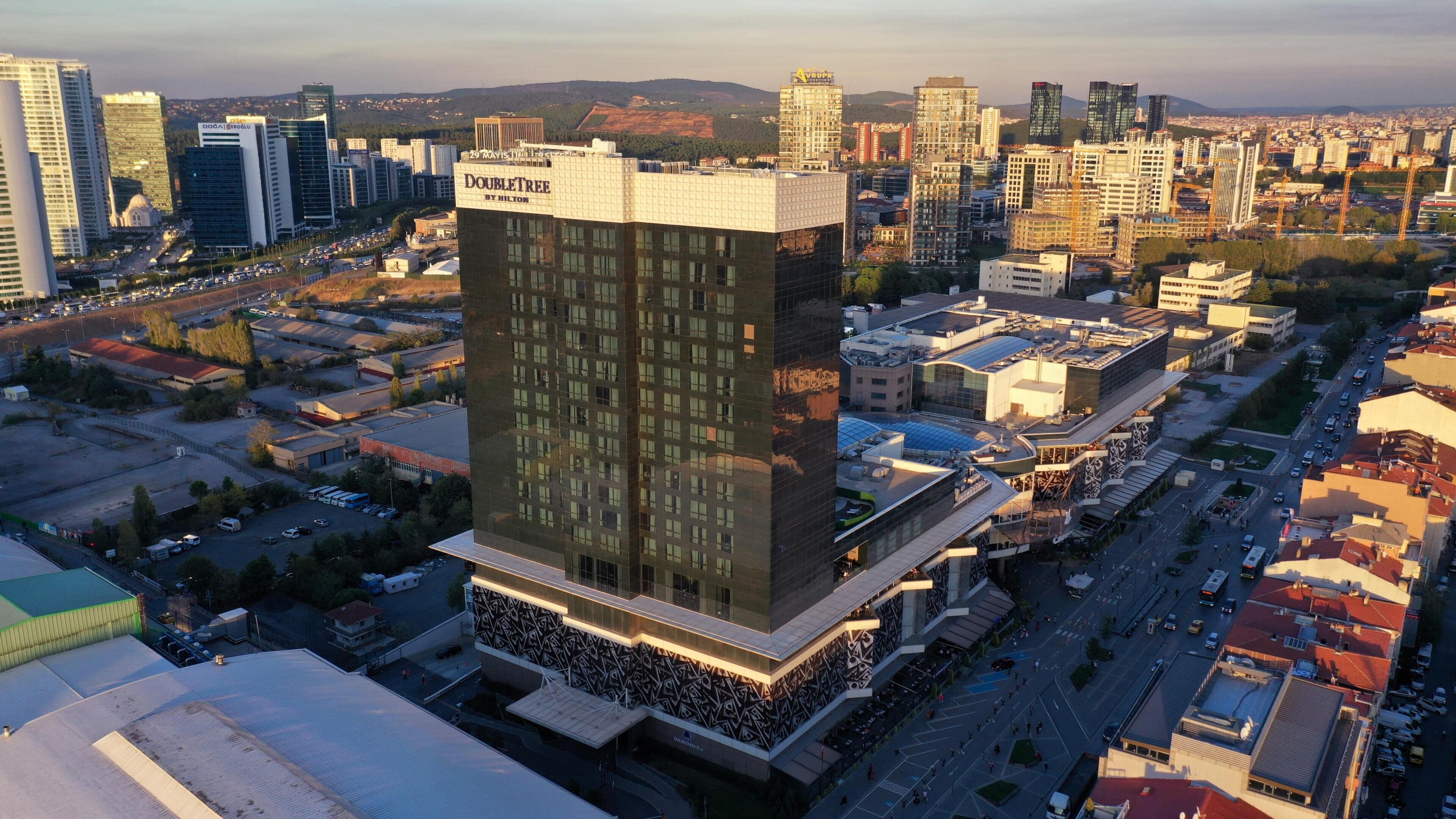 Un hôtel moderne, reconnaissable à son grand panneau « DoubleTree by Hilton », situé au cœur d’un paysage urbain composé de différents bâtiments et bordé d’une rue animée.