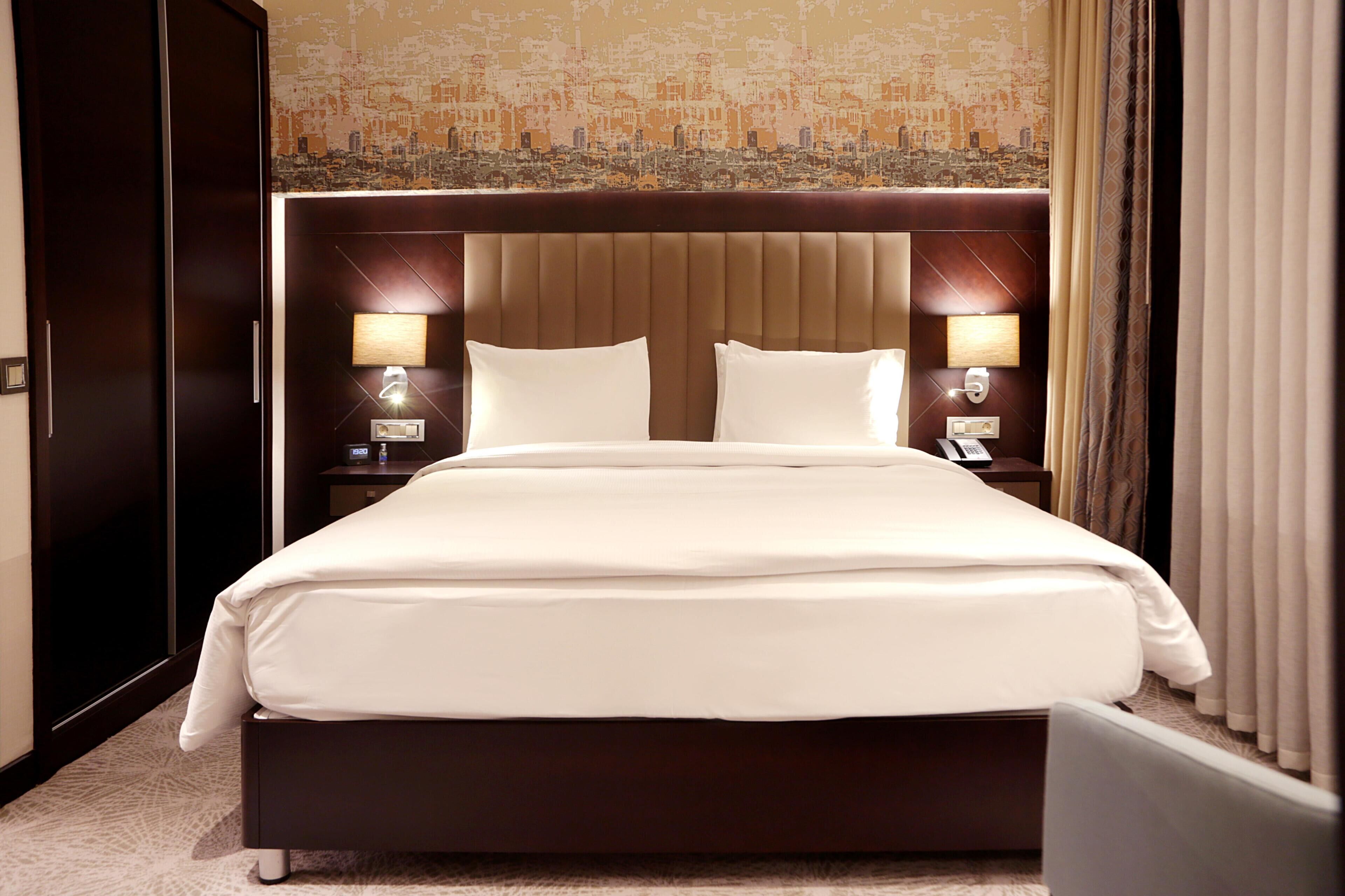 Une chambre d’hôtel avec un grand lit, deux tables de chevet avec des lampes, une tête de lit ornée d’une fresque représentant un paysage urbain et une armoire.