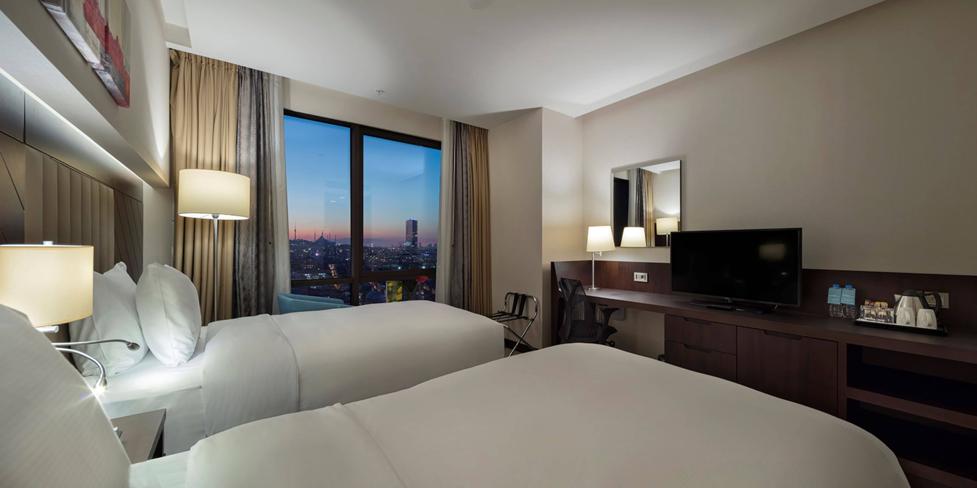 A hotel room with two beds, a desk, a TV, and a city view.
