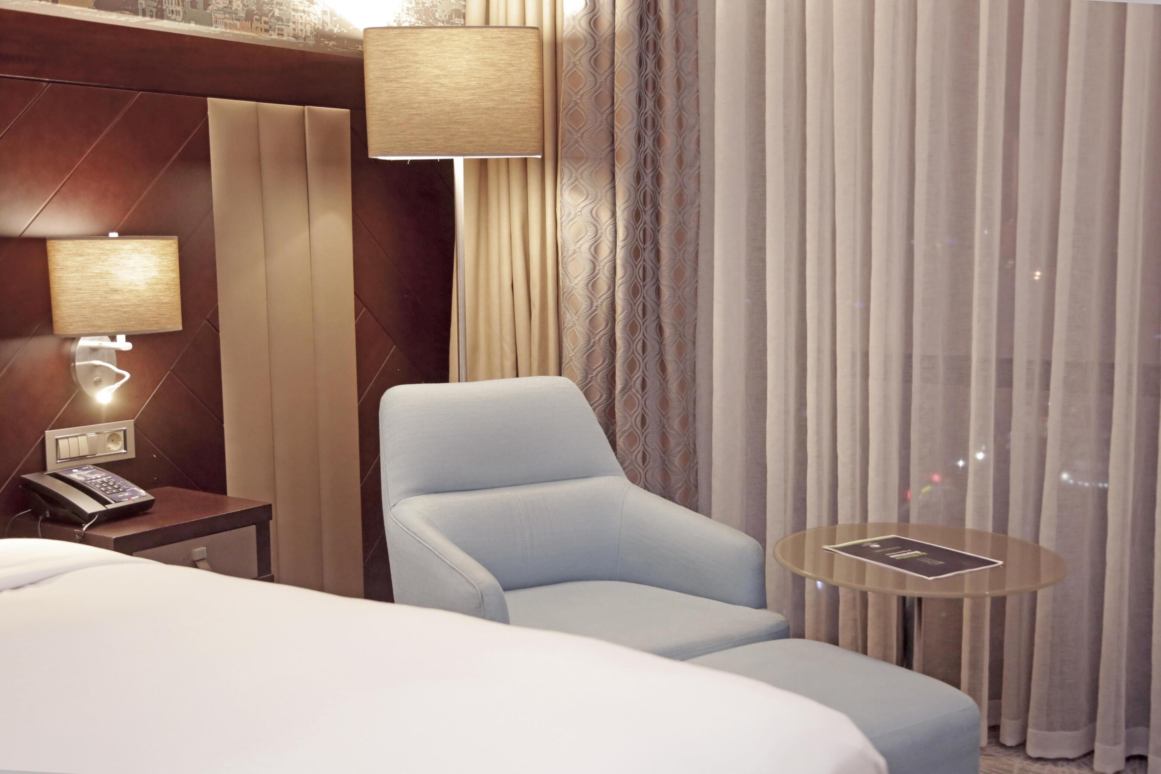 A hotel room with a bed, a chair, a small table, a lamp, and a telephone on a nightstand.