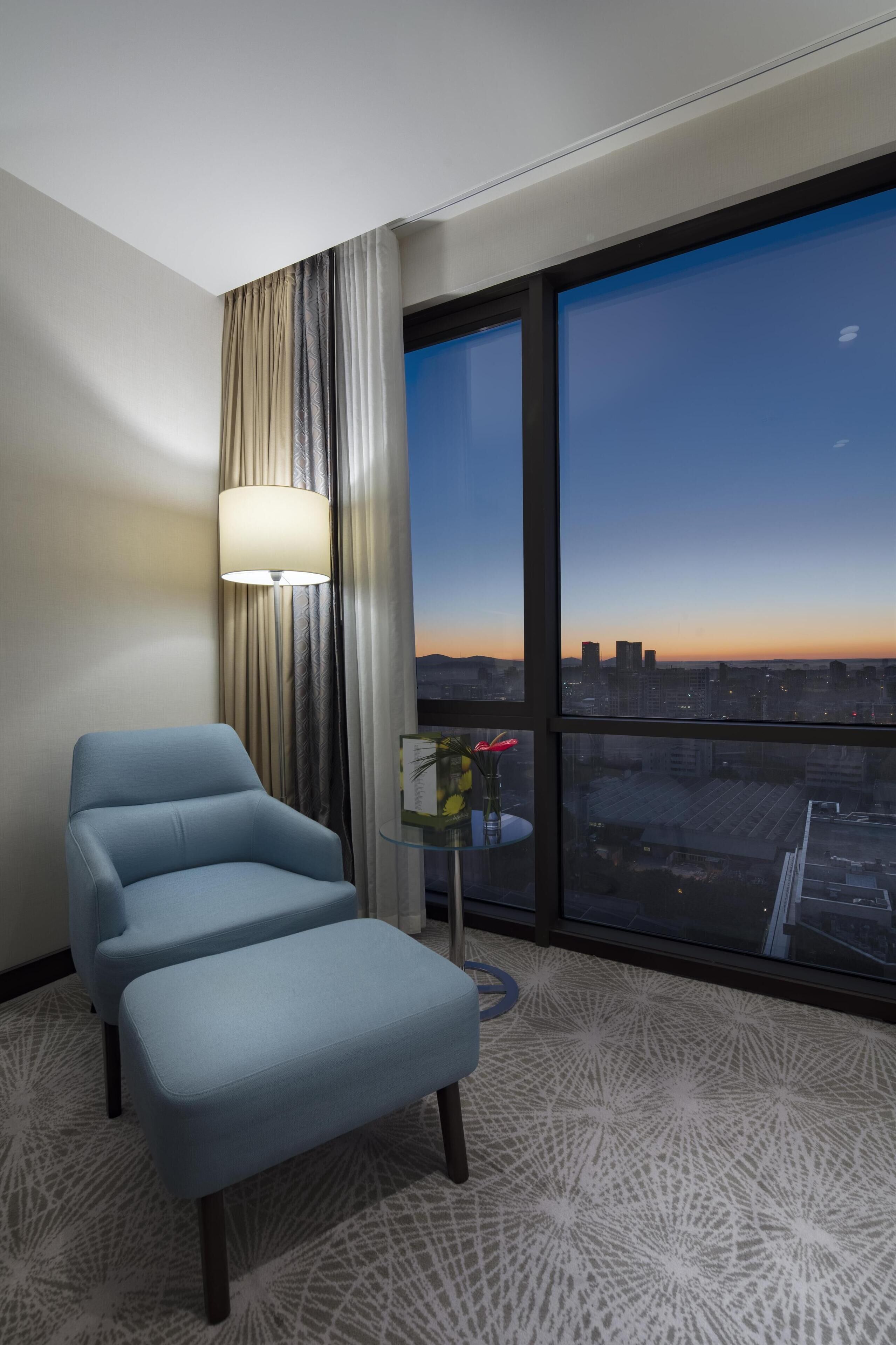 Habitación de hotel con un ventanal grande, un sillón azul, una mesita con un florero y una alfombra estampada.