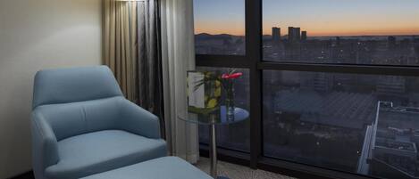 Habitación de hotel con un ventanal grande, un sillón azul, una mesita con un florero y una alfombra estampada.