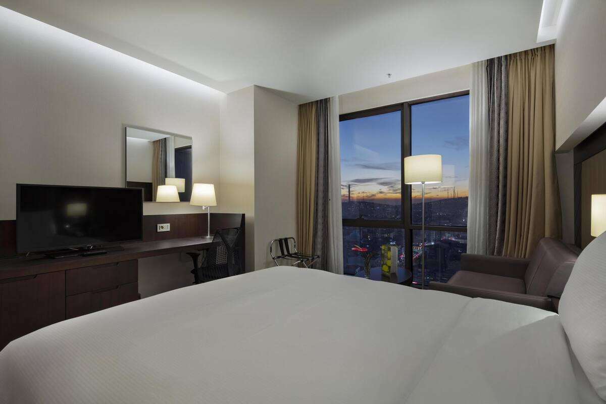 A hotel room with a bed, a desk with a television, a chair, a large window with curtains, and a view of a cityscape at dusk.