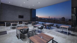 Ein Dachterrassen-Essbereich mit Tischen und Stühlen, Blick auf die Stadt und einem an der Wand befestigten Flachbildfernseher.