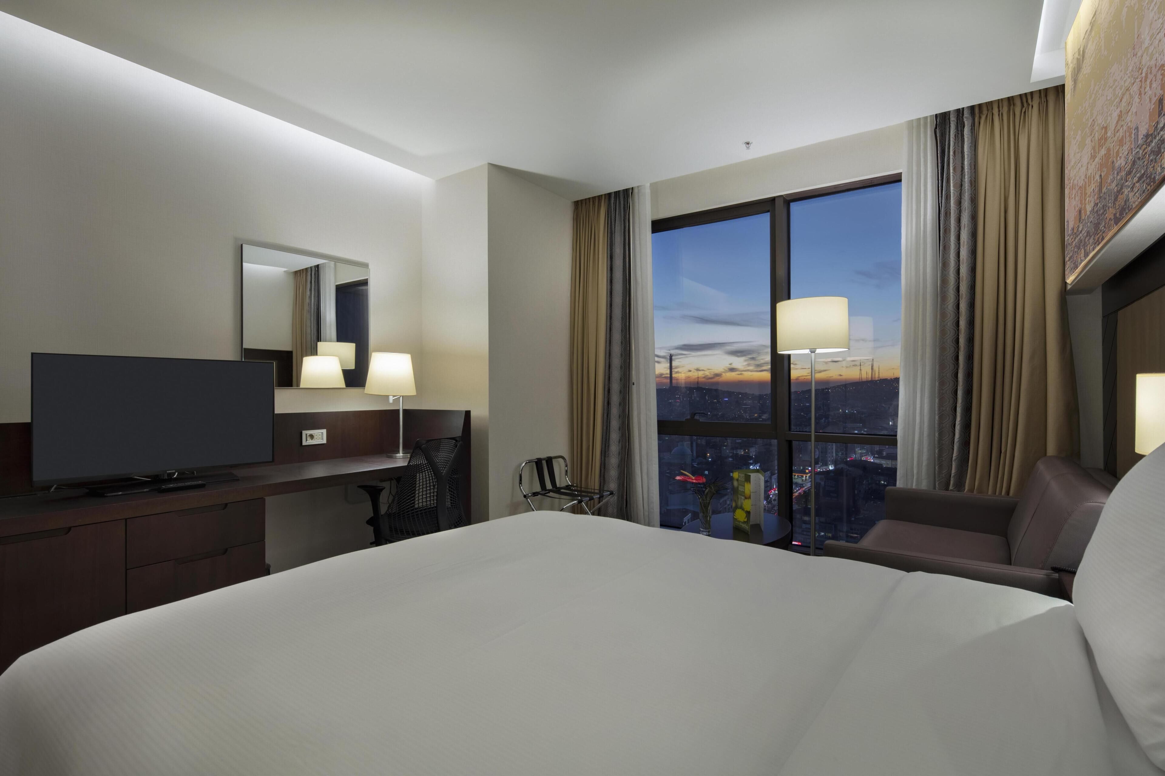 A hotel room with a large bed, a desk with a flat-screen TV, a chair, a window with a city view, and a painting on the wall.