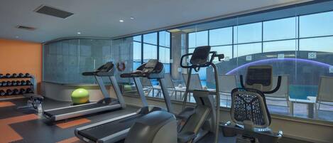 Une salle de sport équipée de tapis de course, d’un vélo elliptique et d’un rameur, avec une grande fenêtre donnant sur un immeuble.