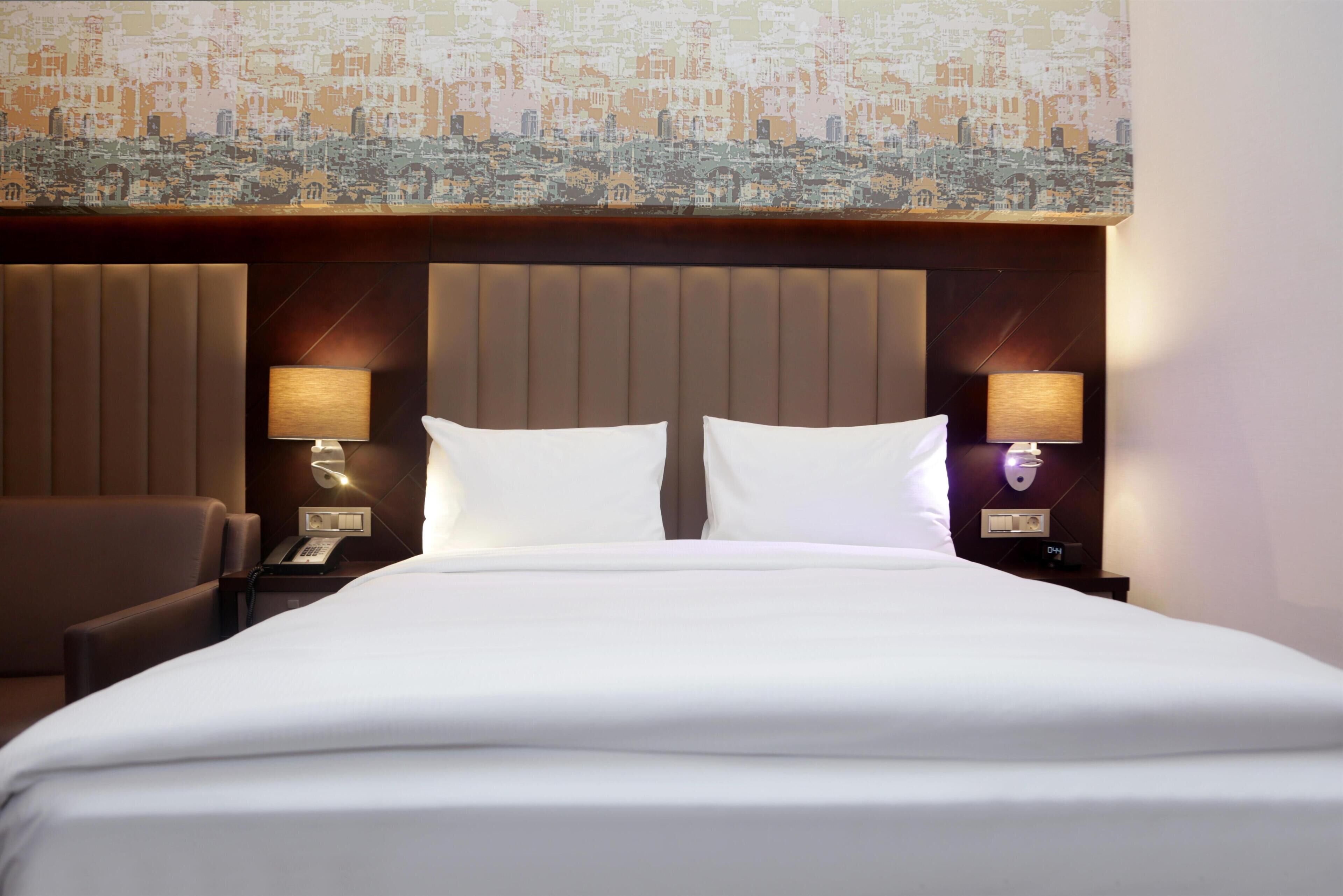 Una habitación de hotel con una cama grande, dos lámparas de noche, una silla y un mural con vista a la ciudad en la pared.