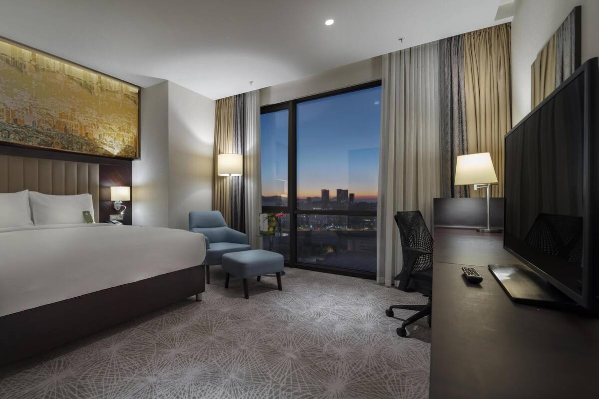 A hotel room with a large bed, a desk with a TV, a chair, and a view of the city at dusk.