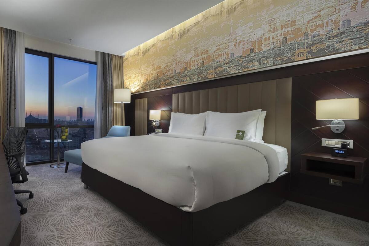 A hotel room with a large bed, a desk with a TV, a chair, a small table, and a view of the city through large windows.