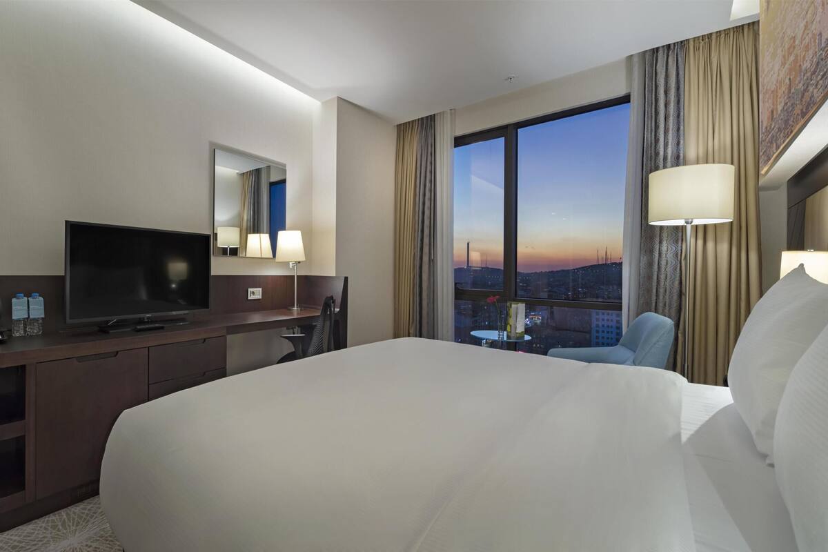 A hotel room with a large bed, a desk with a TV, a chair, and a view of the cityscape at sunset.