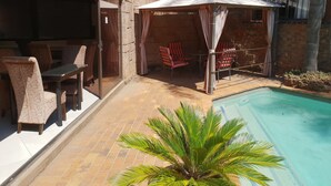 Un espace aménagé au bord de la piscine, avec un coin salon couvert, des chaises et un palmier.
