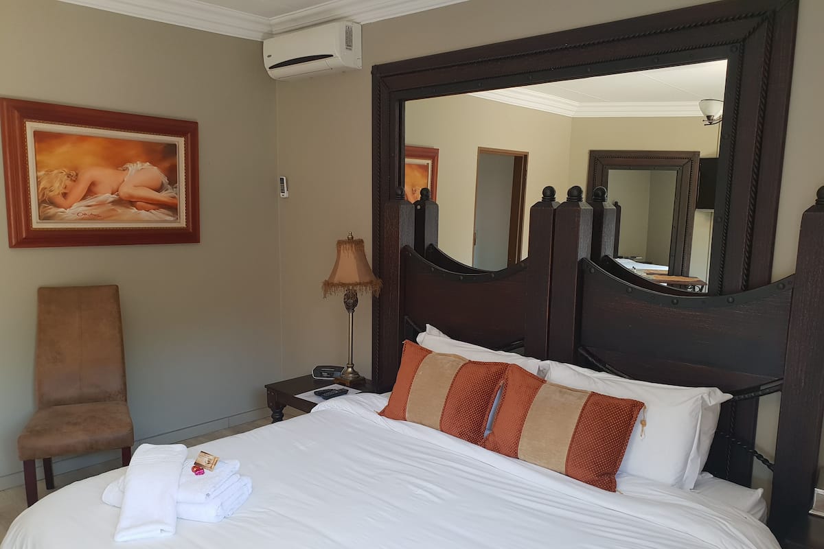 A neatly made bed with white linens and brown striped pillows, a wooden nightstand with a lamp, a framed painting on the wall, and a large mirror above the bed.