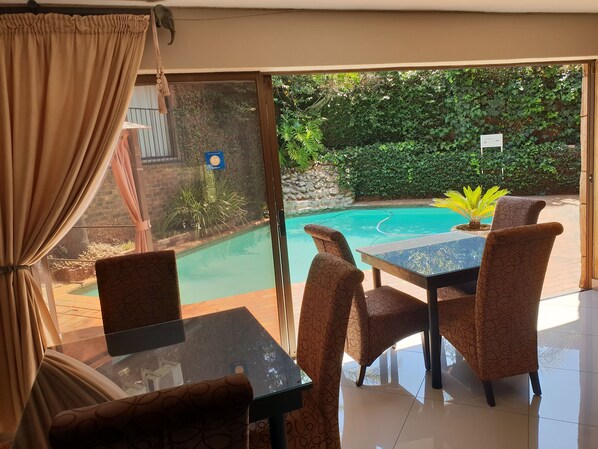 A room with a glass door leading to a pool, a table with chairs, and a view of greenery outside.