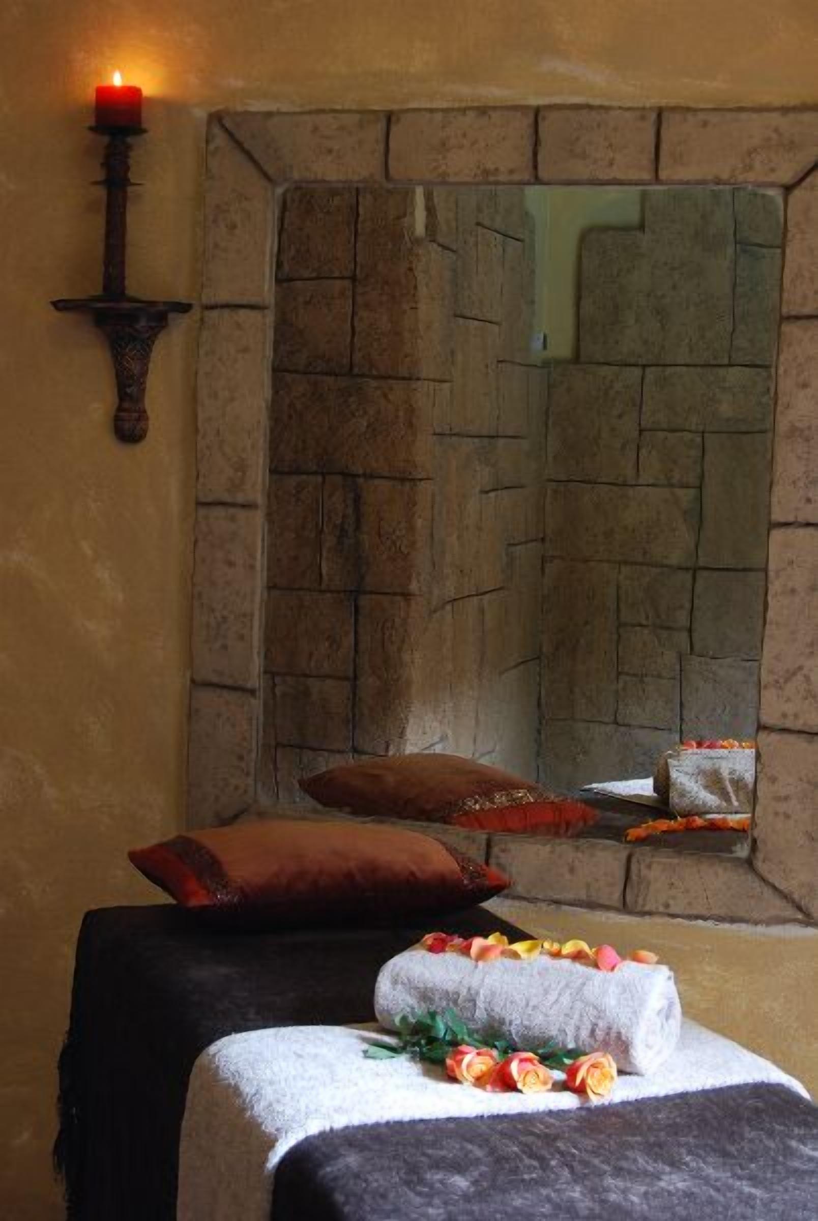A room with a stone fireplace, a candle on a wall-mounted holder, a bed with a dark cover, and a towel with flowers.