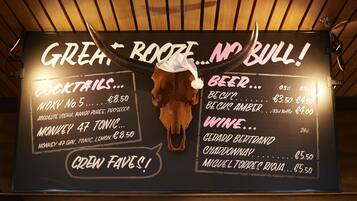 Un cartel de menú con techo de madera y una decoración de calavera. Ofrece cócteles, cerveza y vino con sus precios.