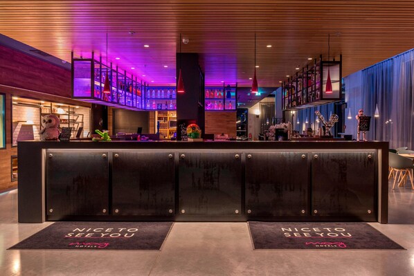 Zona de recepción de un hotel con diseño moderno, que incluye un bar con diferentes botellas, un peluche y un cartel que dice 'Nice to see you'.
