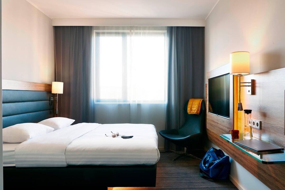 A hotel room with a bed, a desk with a laptop, a chair, a TV, and a lamp.