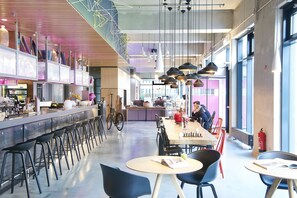 Un café moderno con una barra larga, taburetes y mesas con sillas. Hay bicicletas aparcadas contra la pared, y un extintor es visible en la pared.