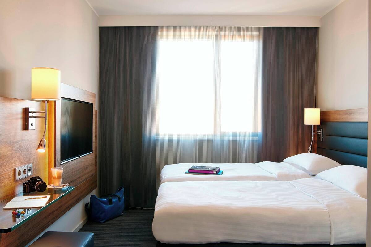 A hotel room with a bed, a desk with a lamp, a TV, and a window with curtains.