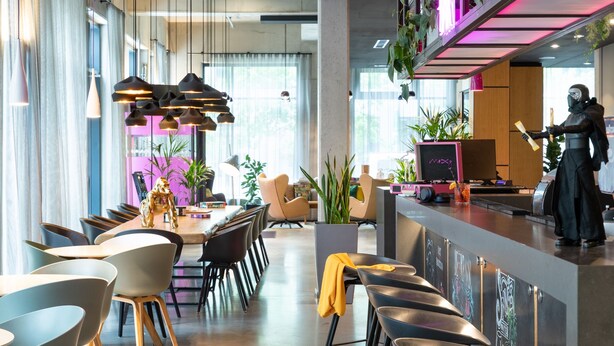 Un restaurant moderne doté d’une longue table à manger, de nombreuses chaises, de luminaires suspendus et d’un bar avec des tabourets.