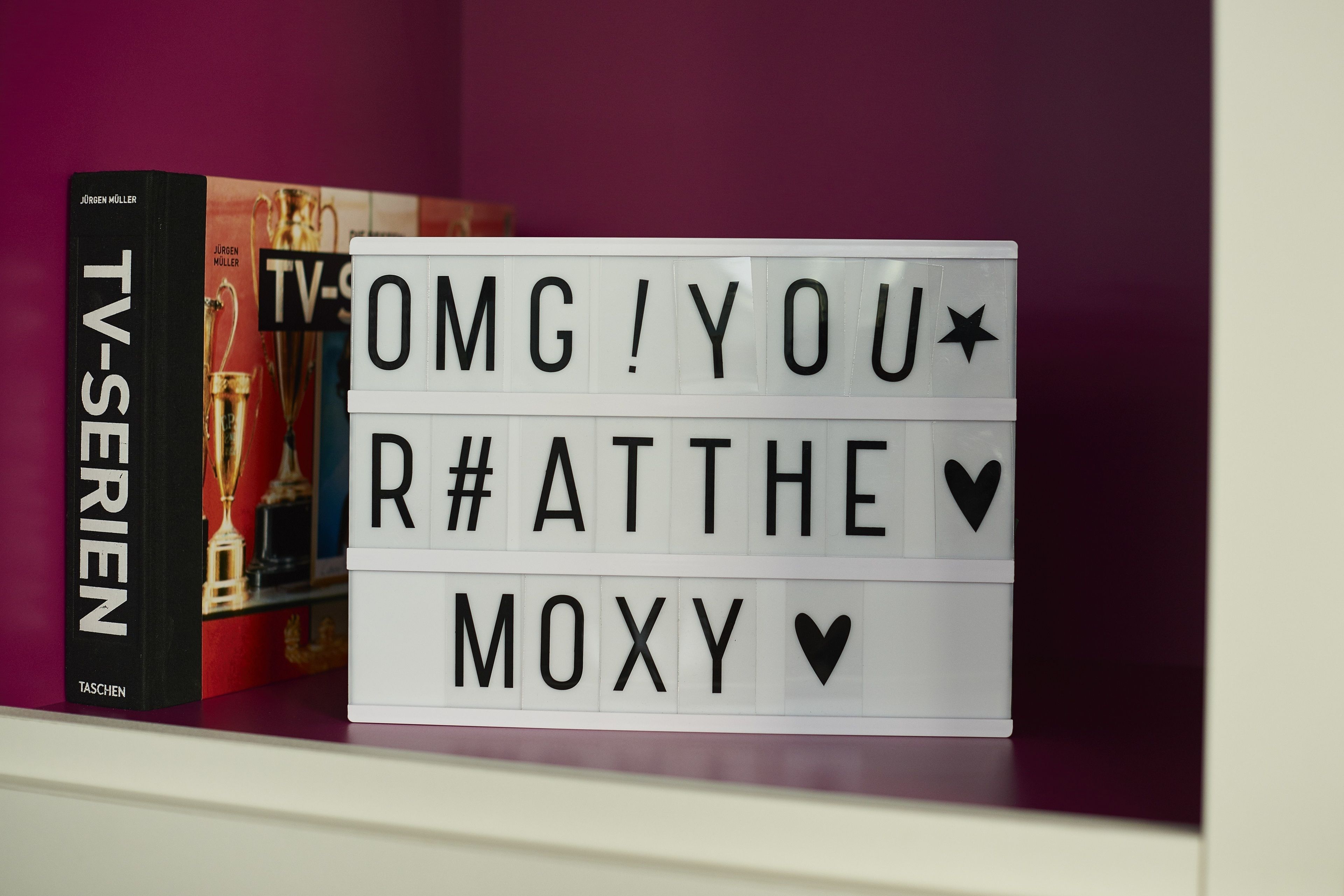 Una estantería con un libro titulado "TV-Serien" y una luz caja con el texto "OMG! YOU R# AT THE MOXY V".