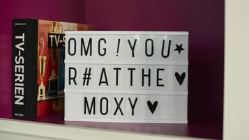 Una estantería con un libro titulado "TV-Serien" y una luz caja con el texto "OMG! YOU R# AT THE MOXY V".
