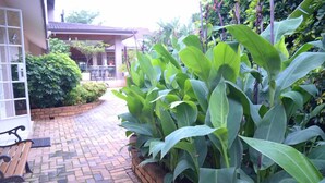 A brick pathway leading to a covered patio area with lush green plants and a bench. - Ah Ha Guest House (Germiston)