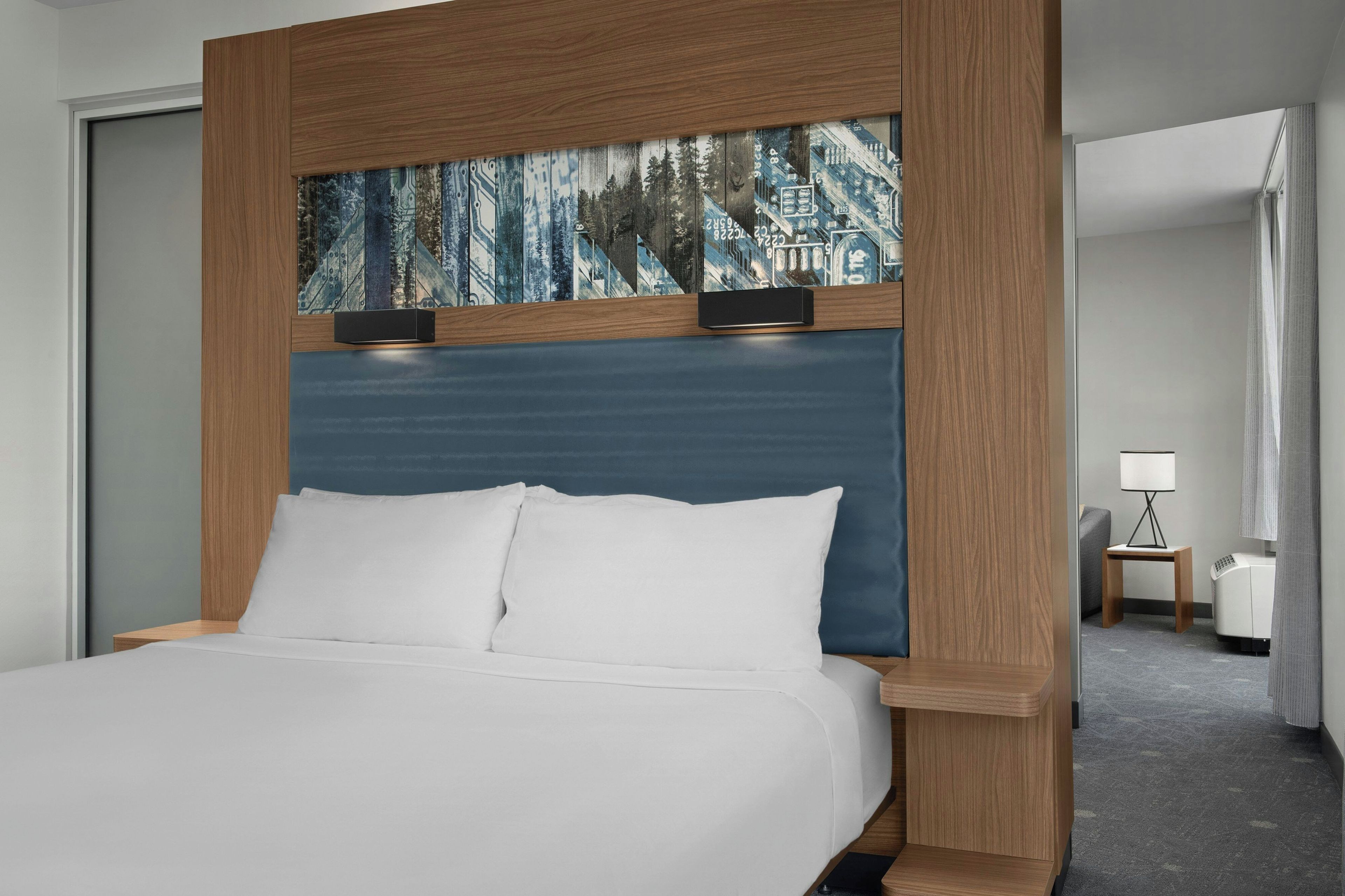 A hotel room with a bed, a headboard with a blue panel, and a nightstand with a lamp.