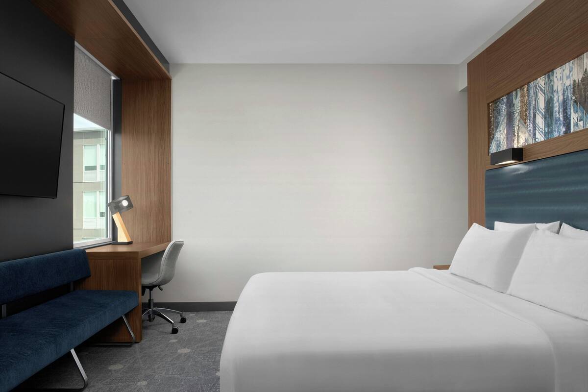 A hotel room with a bed, a desk with a chair, a television, and a painting on the wall.