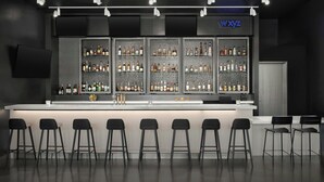Un'area bar moderna con un lungo bancone, numerose scaffalature piene di bottiglie e diversi sgabelli da bar neri.