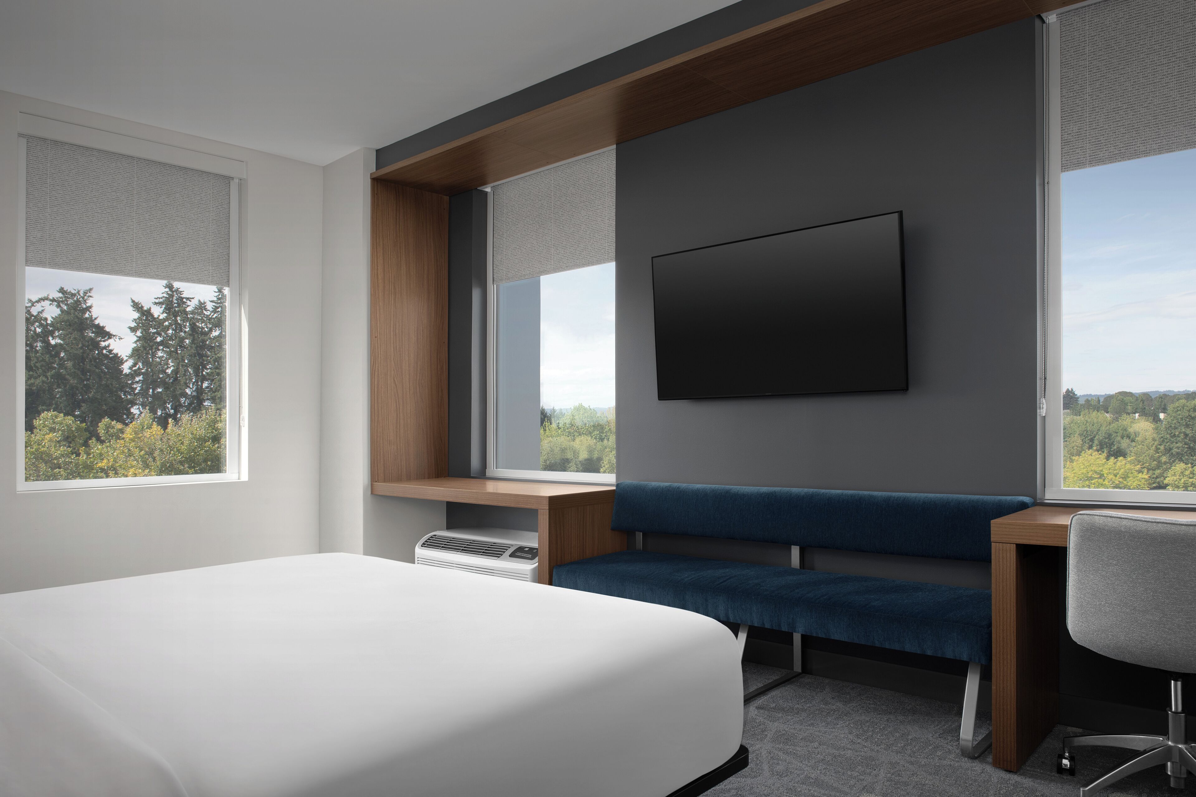 A modern hotel room with a large bed, a desk with a chair, a blue sofa, a flat-screen TV mounted on the wall, and large windows offering a view of trees.