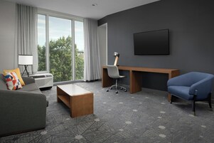 Una habitación de hotel moderna con un ventanal grande, un escritorio, un sillón azul, un sofá gris y una alfombra estampada.