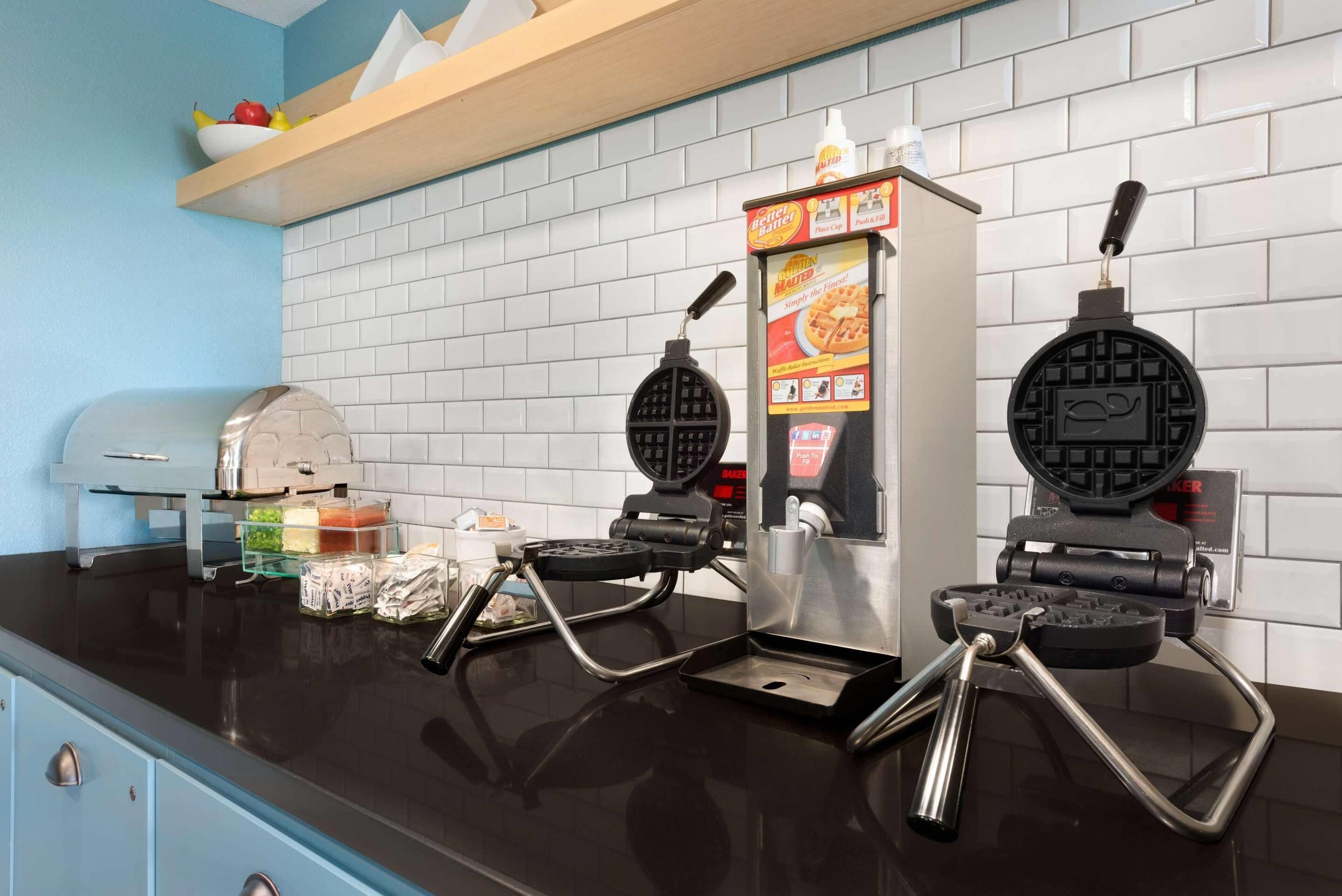 A kitchen with two waffle makers, a toaster, and a shelf with fruits.