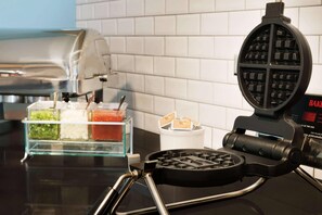 A waffle maker on a kitchen counter with a glass container of condiments and a white container with napkins.
