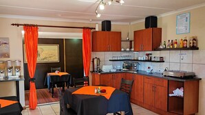 A compact kitchen with wooden cabinets, a microwave, and a coffee maker. There are two tables with orange tablecloths, and a dining area with chairs. - Premiere Guest House (Bloemfontein)