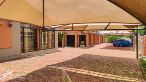 A covered outdoor area with a paved walkway, a car parked under a canopy, and a building with large windows and a sign reading "Premier." - Premiere Guest House (Bloemfontein)