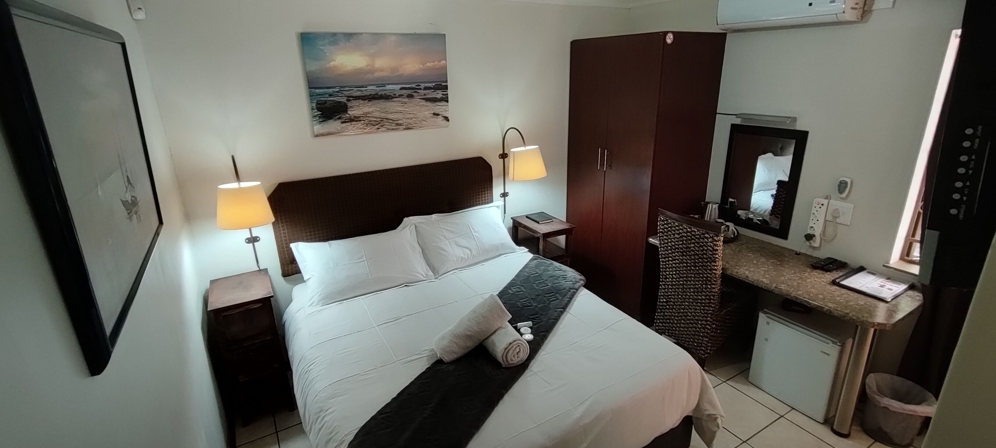 Une chambre d’hôtel compacte comprenant un lit, des lampes de chevet, un bureau avec un ordinateur, une armoire et une petite kitchenette.