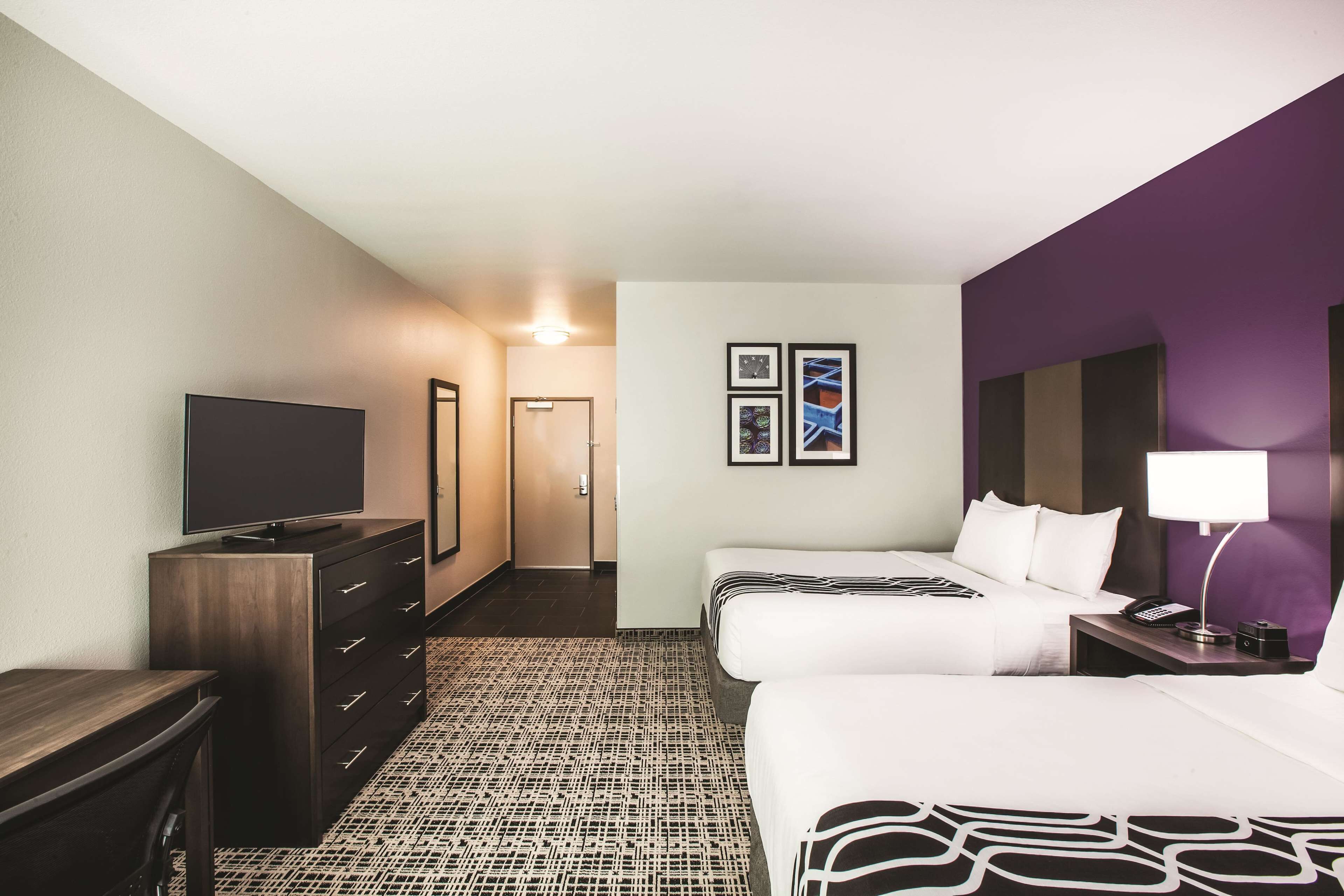 A hotel room with a large flat-screen TV, a dresser, two beds with white bedding, a patterned carpet, and a purple accent wall.