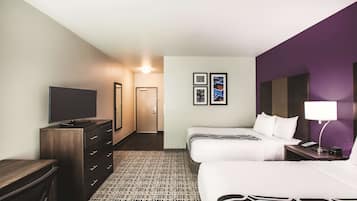 A hotel room with a large flat-screen TV, a dresser, two beds with white bedding, a patterned carpet, and a purple accent wall.