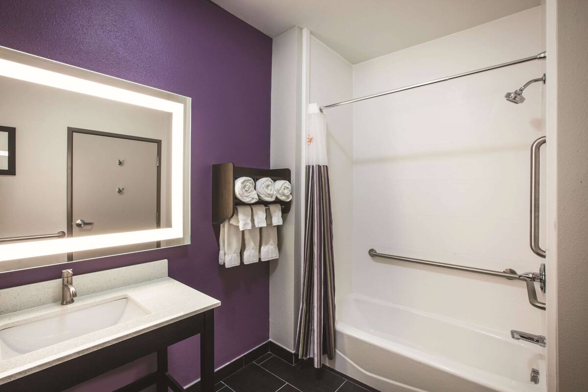 Een badkamer met een wit wastafel, een spiegel, een douche met een handgreep en handdoeken aan een rek.