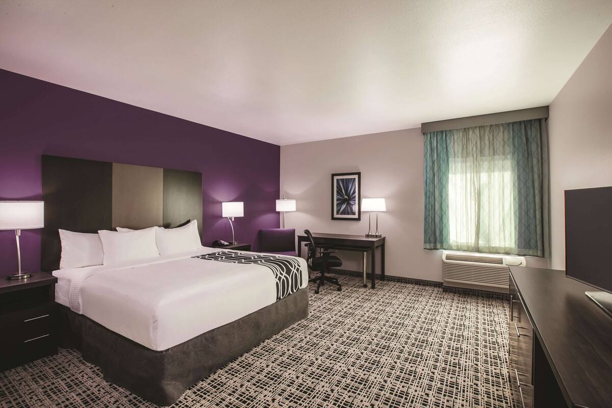 Een hotelkamer met een groot bed, twee nachtkastjes met lampen, een bureau met een stoel, een televisie en een raam met gordijnen.