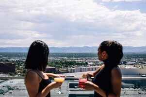 Dos personas de pie en una azotea, con un paisaje urbano y montañas al fondo.