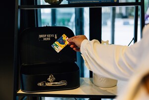 Una mano depositando una tarjeta en una caja negra con la etiqueta 'DROP KEYS HERE'.
