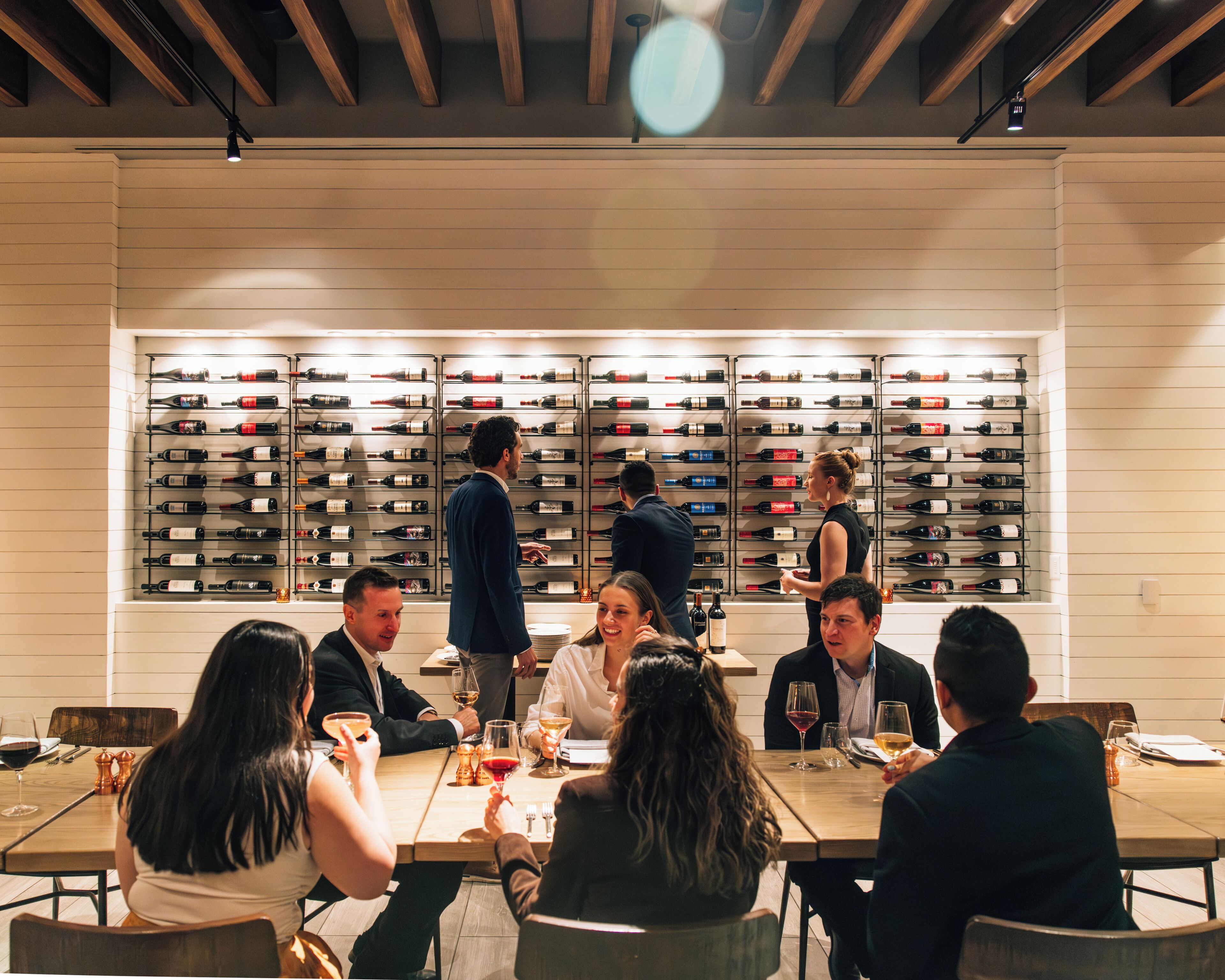 A group of people seated at a table in a restaurant with a wine rack in the background.