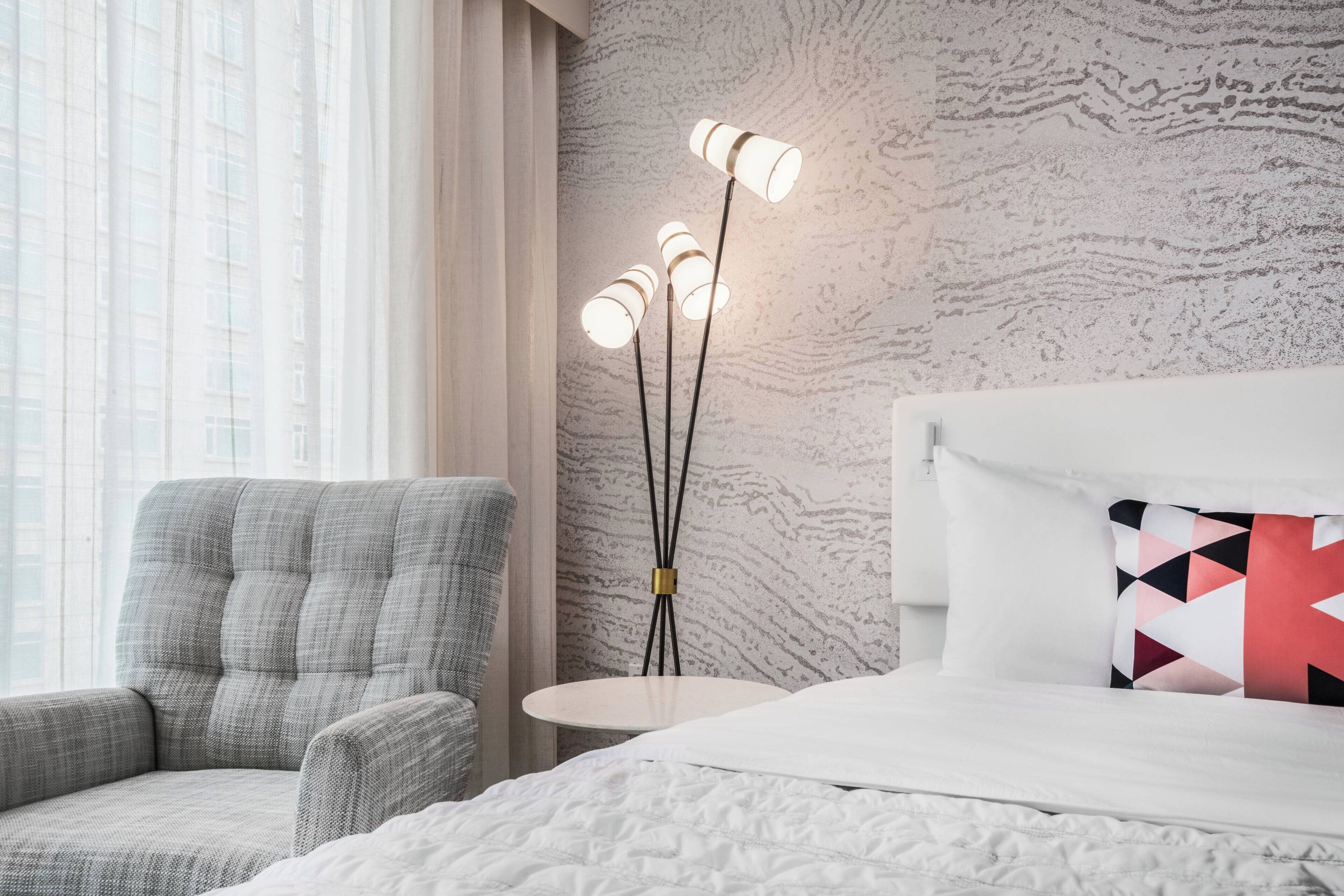 Une chambre d’hôtel avec un lit, une chaise, une petite table et une lampe sur pied.
