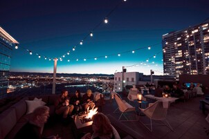 Un bar en la azotea con guirnaldas de luces, mobiliario exterior y vistas a la ciudad al anochecer.