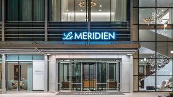 A modern hotel entrance with a neon sign reading "Le MERIDIEN" and a visible reception area.