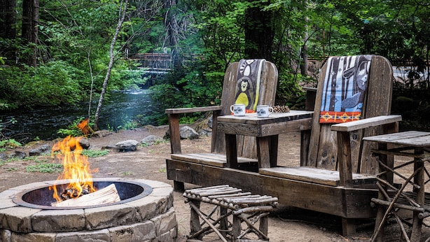A campfire, wooden chairs, and a bench in a forested area with a view of a bridge. - Union Creek Resort (Prospect)