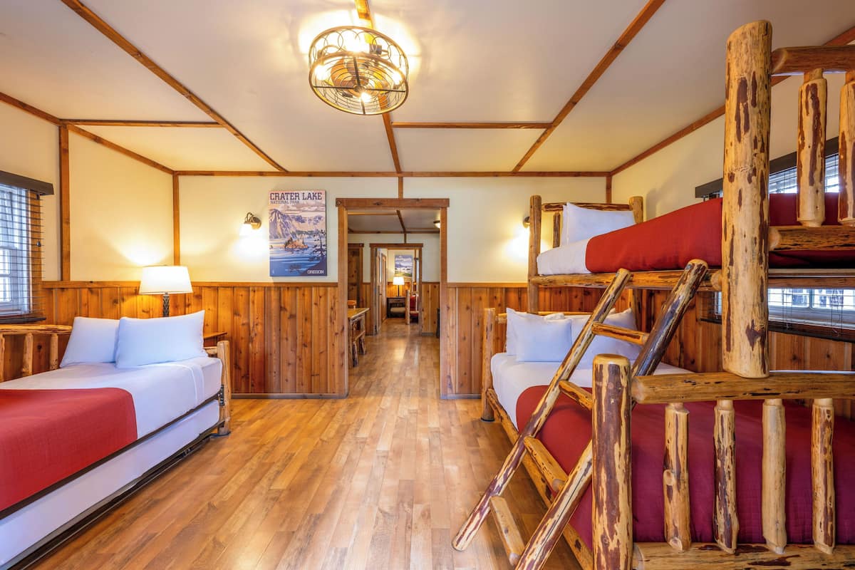 Quarto estilo cabana com paredes e chão de madeira, uma cama beliche, uma escada, um cartaz na parede e um corredor que dá para outro quarto.